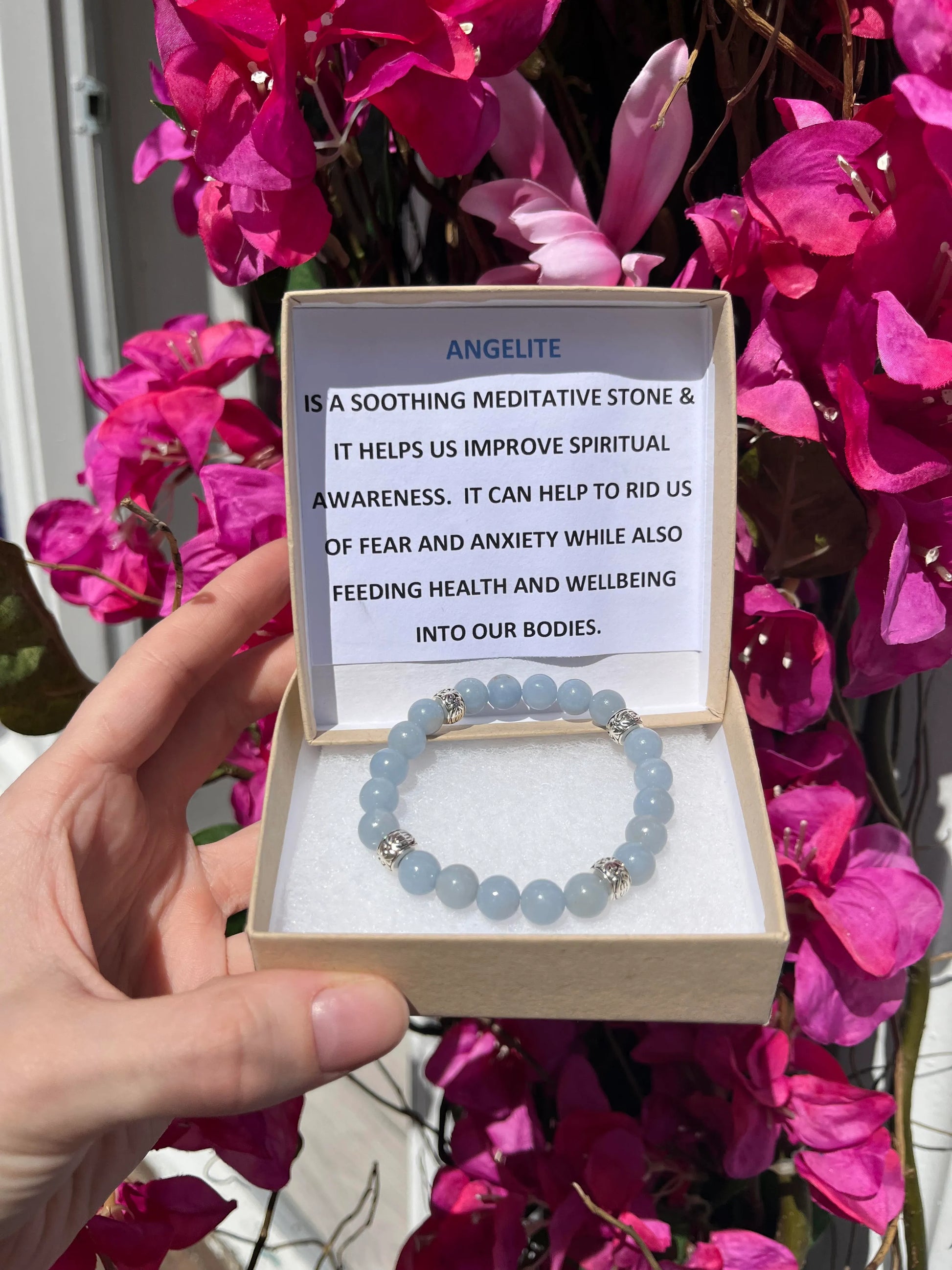 A crystal bracelet in a brown box with a typed description in the lid that reads: Angelite is a soothing meditative stone & helps us improve spiritual awareness. It can help to rid of us fear and anxiety while also feeding health and wellbeing into our bodies.